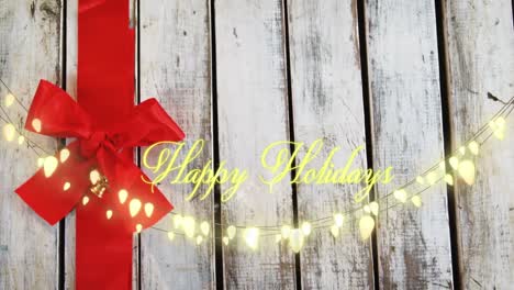 Animation-of-text,-happy-holidays,-in-yellow,-over-string-lights-and-red-bow-on-painted-wood-boards