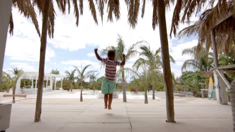 happy caucasian boy running and raising hands at swimming pool at beach house with palm trees