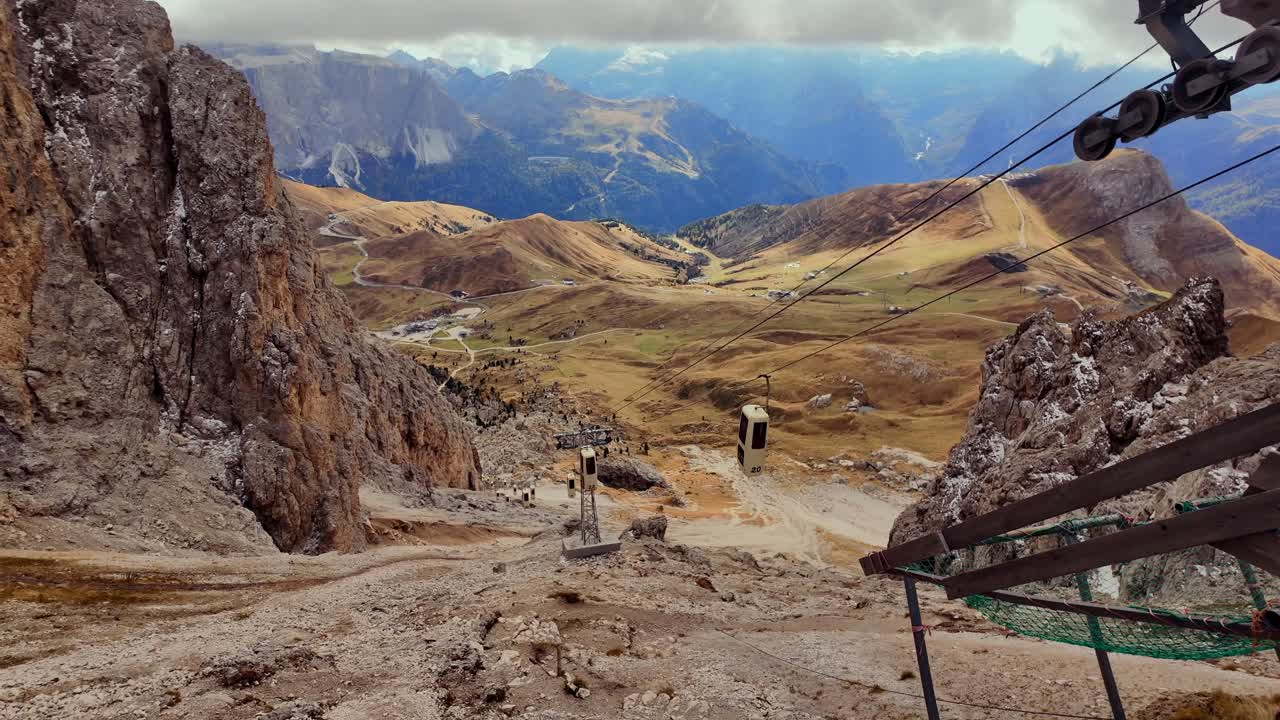 Cable car descends from a high elevation in the Dolomites, with a stunning view of the surrounding rugged mountains and valleys. The shot captures the steep terrain and distant alpine peaks.