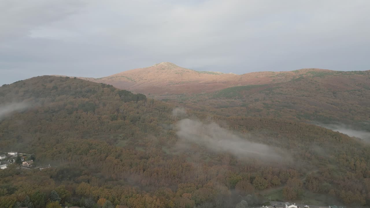 reverse flight with a drone in a mountainous area with a central peak full of chestnut trees and as it is autumn they begin to bring out their colors, buildings and also morning fog appear