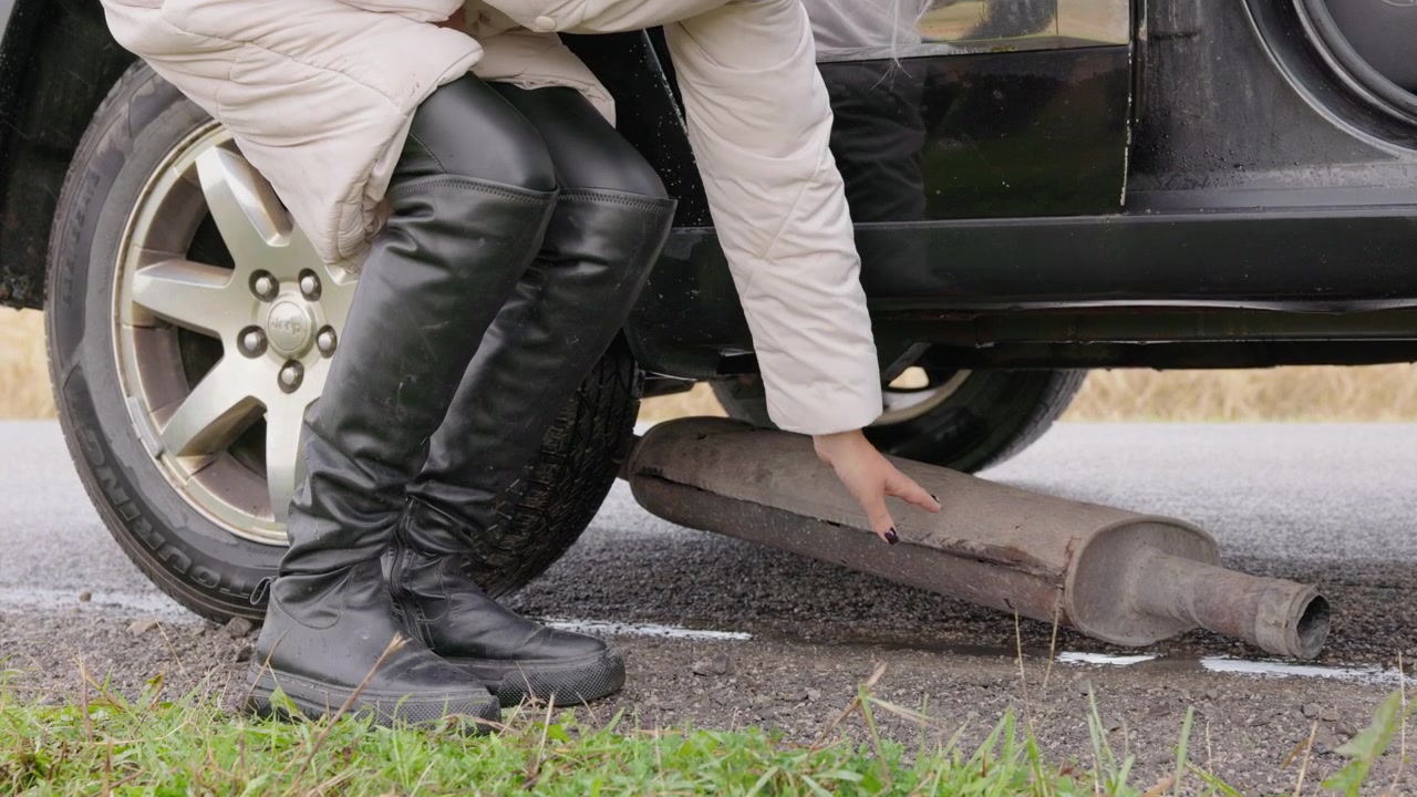 Woman Inspecting Detached Car Exhaust Pipe on Roadside