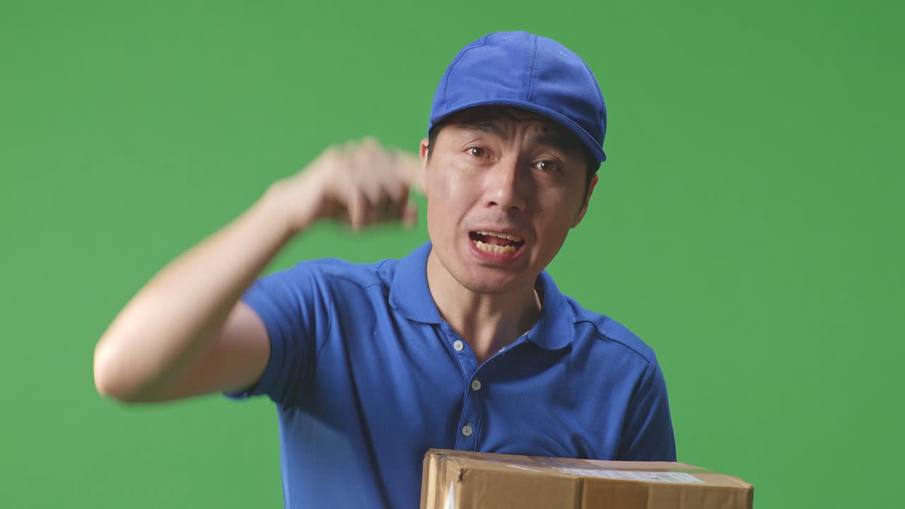 푸른 유니폼을 입은 불만족한 아시아 남성 택배인이 스튜디오의 녹색 스크린 배경에서 상자를 배달하는 동안 카메라에 소리치고 있습니다.