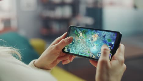 close up on hands playing an rpg strategy video game on a horizontally held smartphone at home living room. feminine hands tapping the mobile phone screen with a colorful game over the internet.