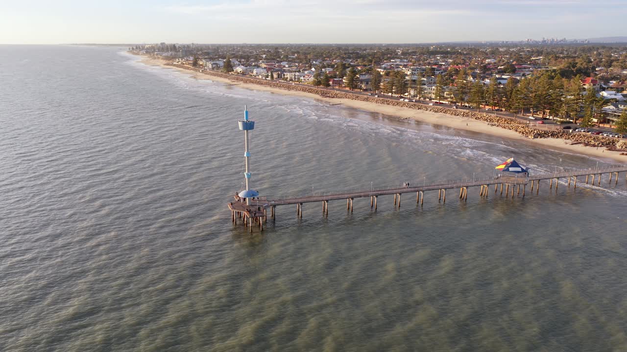 Orbiting drone shot of Brighton Beach jetty at golden hour, with Adelaide's suburbs and city in the background, South Australia