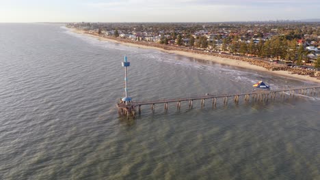orbiting drone shot of brighton beach jetty at golden hour, with adelaide's suburbs and city in the background, south australia