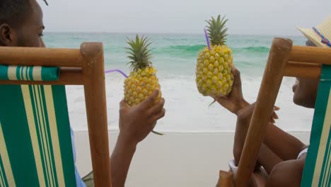 Rückansicht-Eines-Afroamerikanischen-Paares,-Das-Am-Strand-Ananassaft-Toastet,-4k