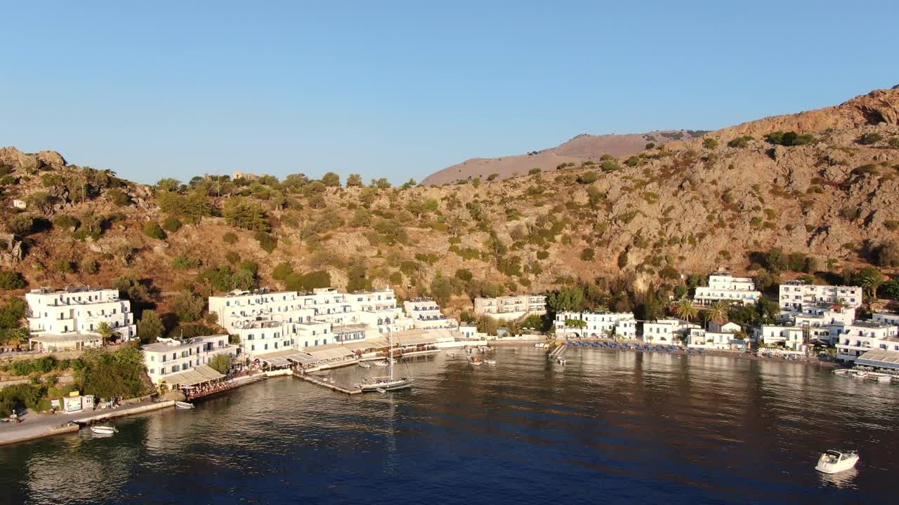Drone view in Greece flying over blue sea in Loutro small white house town and small boats next to a hill on sunset light