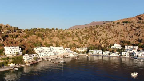 drone view in greece flying over blue sea in loutro small white house town and small boats next to a hill on sunset light