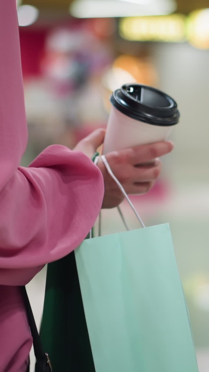 Back view of woman in pink dress holding coffee cup and shopping bags while walking through a brightly lit mall, stores and chairs with tables can be seen with blurred background and mall lights
