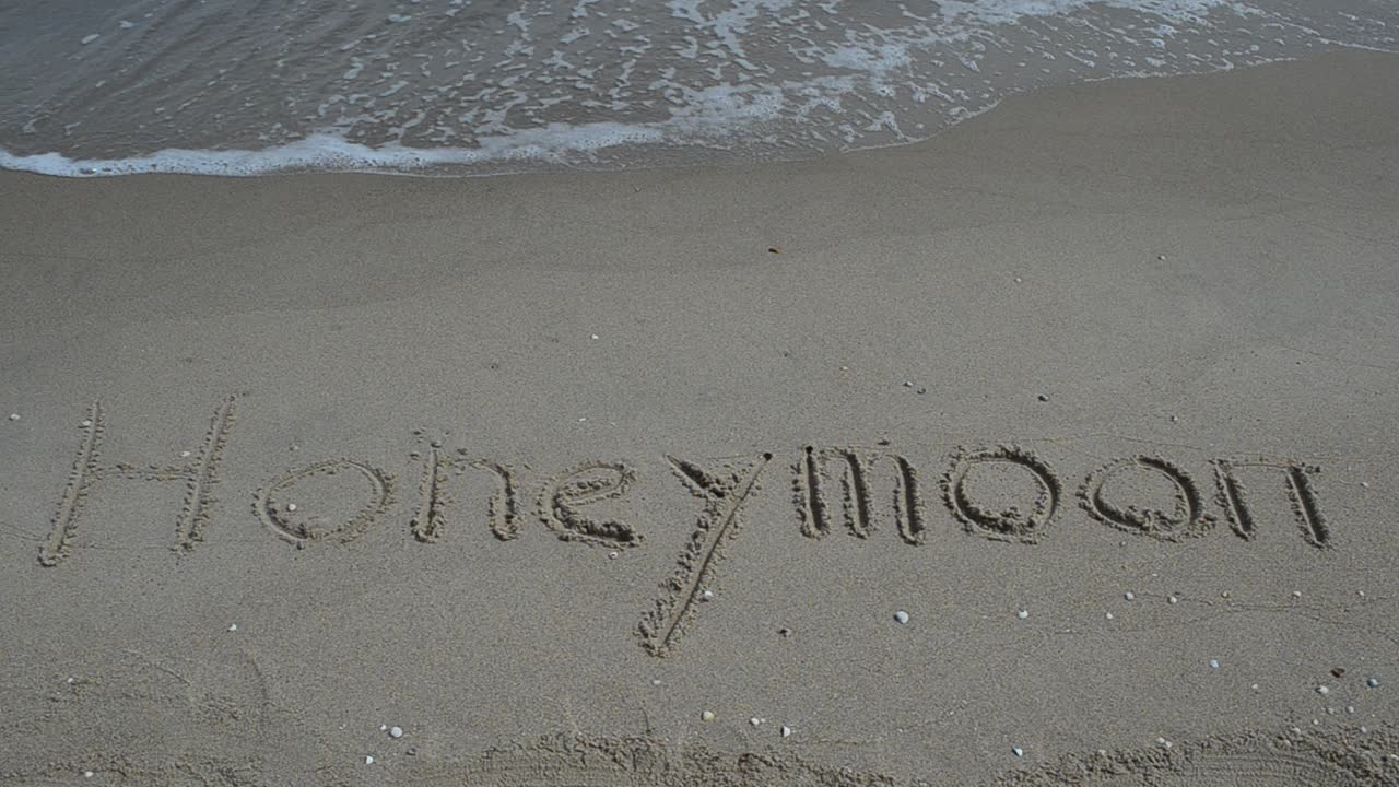 Scritta "Luna di miele" sulla sabbia della spiaggia, con l'onda. Riprese in spiaggia.