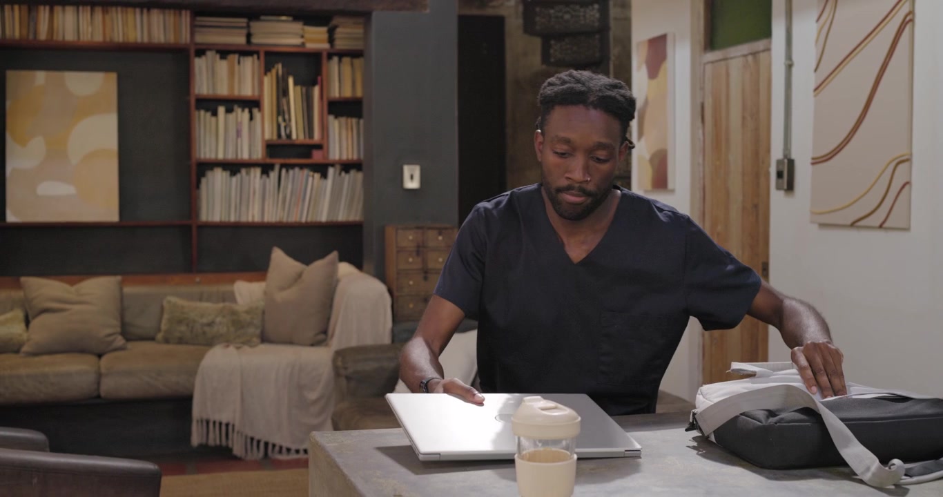 African adult man entering right, wiping island, opening bag, placing laptop, preparing for work