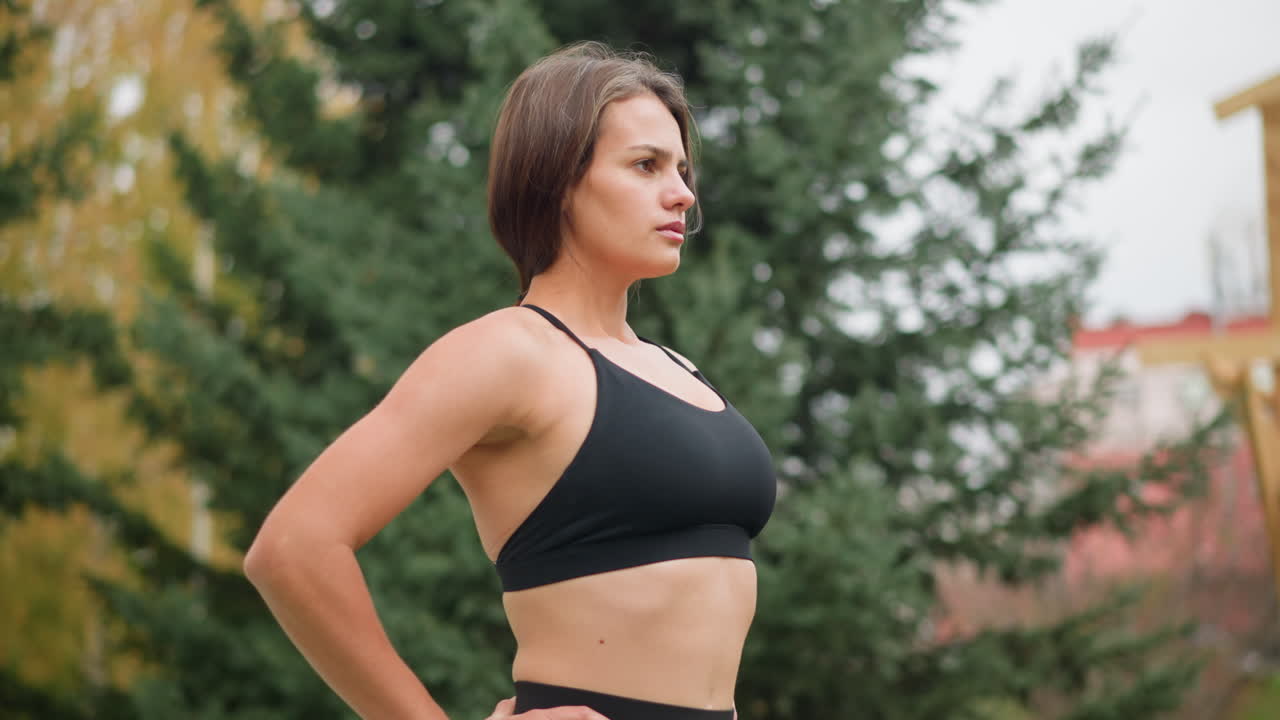 Close-up view of a lady in black sportswear, standing confidently with her hands on her waist and pushing her chest out, showing strength and self-assurance in an outdoor setting