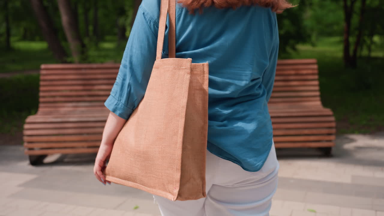 Close up rear view of woman in blue shirt and white pants carrying large brown tote walking toward wooden bench in sunny park, soft light reflecting on pavement