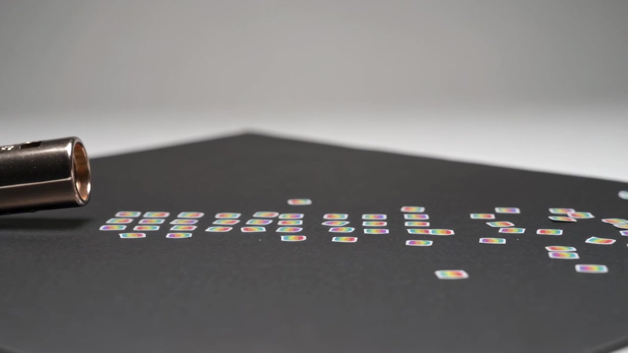 Blowing metal nozzle launching iridescent squares across black mat, scattering into loose rows