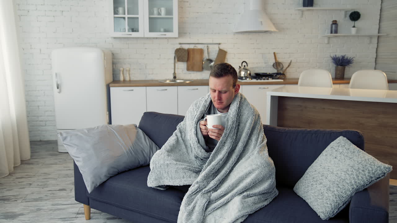 Ill man drinking hot beverage at home. Unwell young man covered with warm blanket sitting on a sofa in the kitchen. Coronavirus pandemic.
