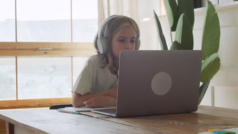 Ein-Hübsches-Und-Intelligentes-Blondes-Mädchen-Mit-Kopfhörern-Macht-Ihre-Hausaufgaben-Und-Schreibt-Auf-Papier,-Während-Sie-Auf-Einen-Laptop-bildschirm-Schaut