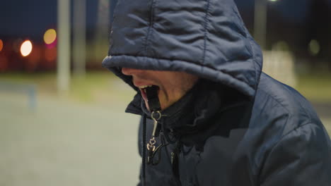 close-up of a man wearing a hood, holding a whistle in his mouth, with a blurred, dimly lit background