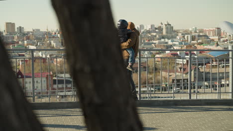 a father holds his young son close to an iron railing as they both gaze out over a cityscape, both are dressed in warm jackets, with view of cars passing by