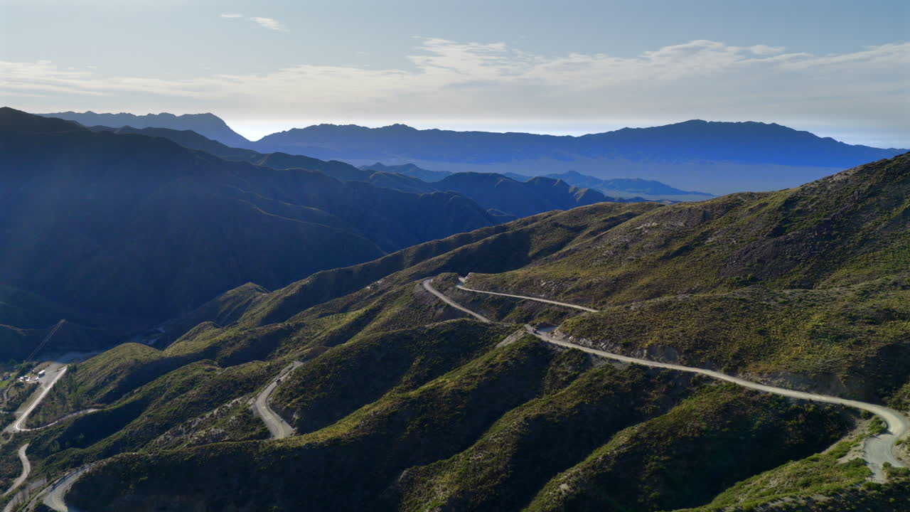 A winding mountain road called Caracoles Villavicencio curves through sunlit green hills, dramatic ridges and blue horizon beyond