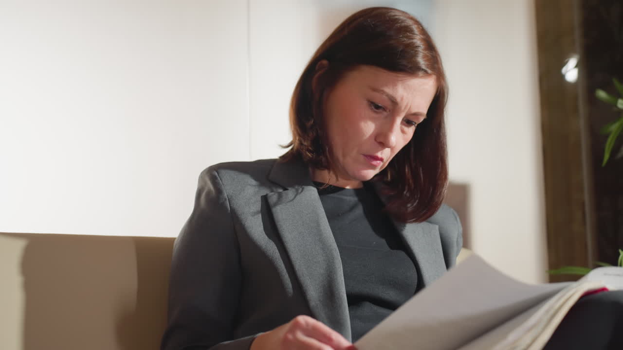 Businesswoman in formal blazer reading important documents with serious focused expression while sitting in modern office setting with natural light and indoor plants in background