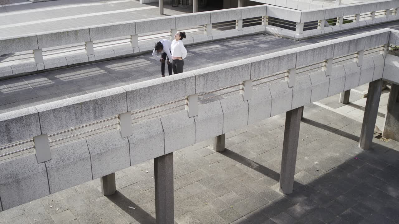 Colegas de trabalho diversos andando em uma ponte, notando um homem na praça que faz uma pausa, pegando um aparelho e apontando.