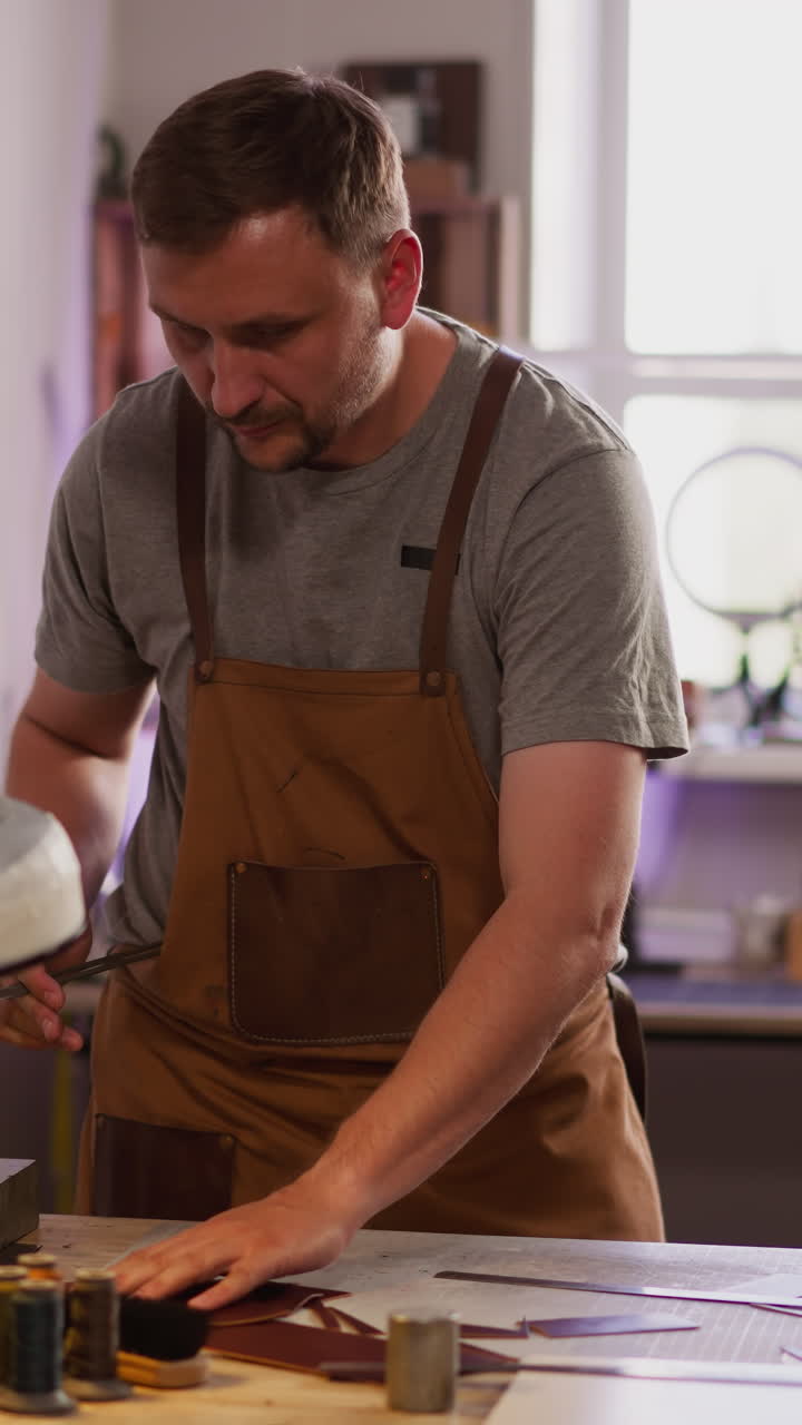Thoughtful man in workwear seeks sharpening tool among leather details on table. Master of traditional industry makes accessory. Skilled tailor at work