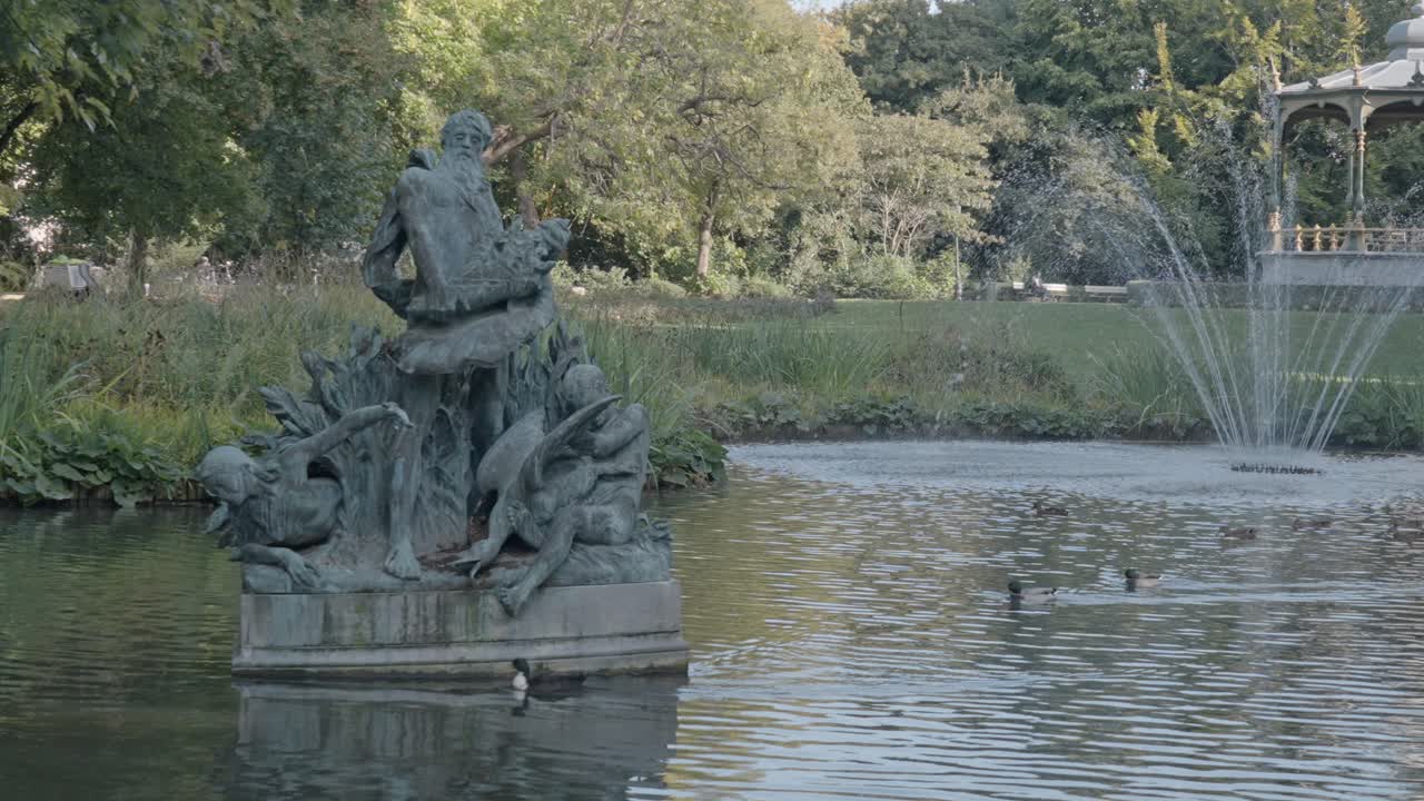 A tranquil view of the Koningin Astridpark in Bruges, Belgium. The scene features a central fountain spraying water and octagonal bandstand standing prominently on the opposite bank