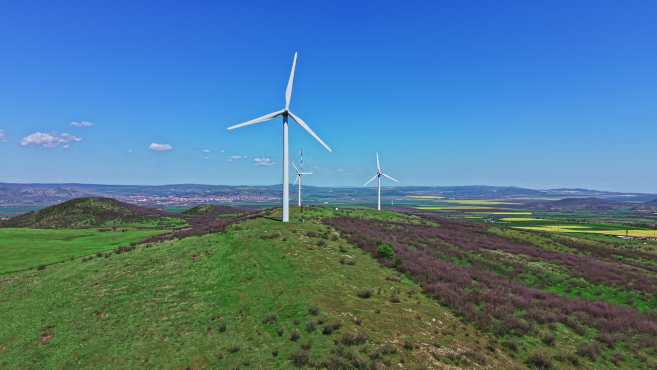 Wind turbines stand tall on a green hill under a bright blue sky in a rural area with fields and small towns in the distance during the day