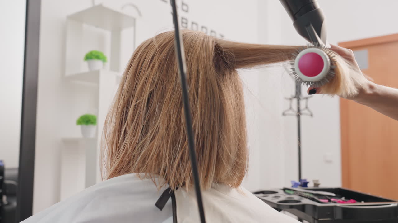 Curly bob receiving professional blowout with dryer, stylist uses round brush and high speed dryer to smooth damp bleached curls, bright modern salon, white cape, product trolley and ring light, quick