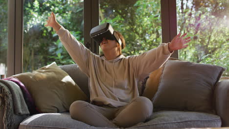 Asiatisches-Mädchen-Lächelt-Und-Gestikuliert,-Während-Es-Ein-VR-Headset-Trägt-Und-Zu-Hause-Auf-Der-Couch-Sitzt