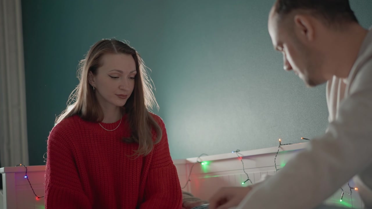 Young woman sits on bed in oversized red sweater while companion leans close beside her, reaching out with gentle support as warm bedroom lighting creates calm, intimate mood in cozy home interior