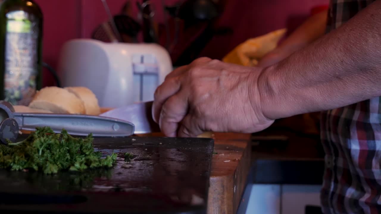 toma manual de manos de ancianas cortando pan con un cuchillo en una vieja cocina con albahaca, aceite de oliva y una tostadora blanca en el fondo