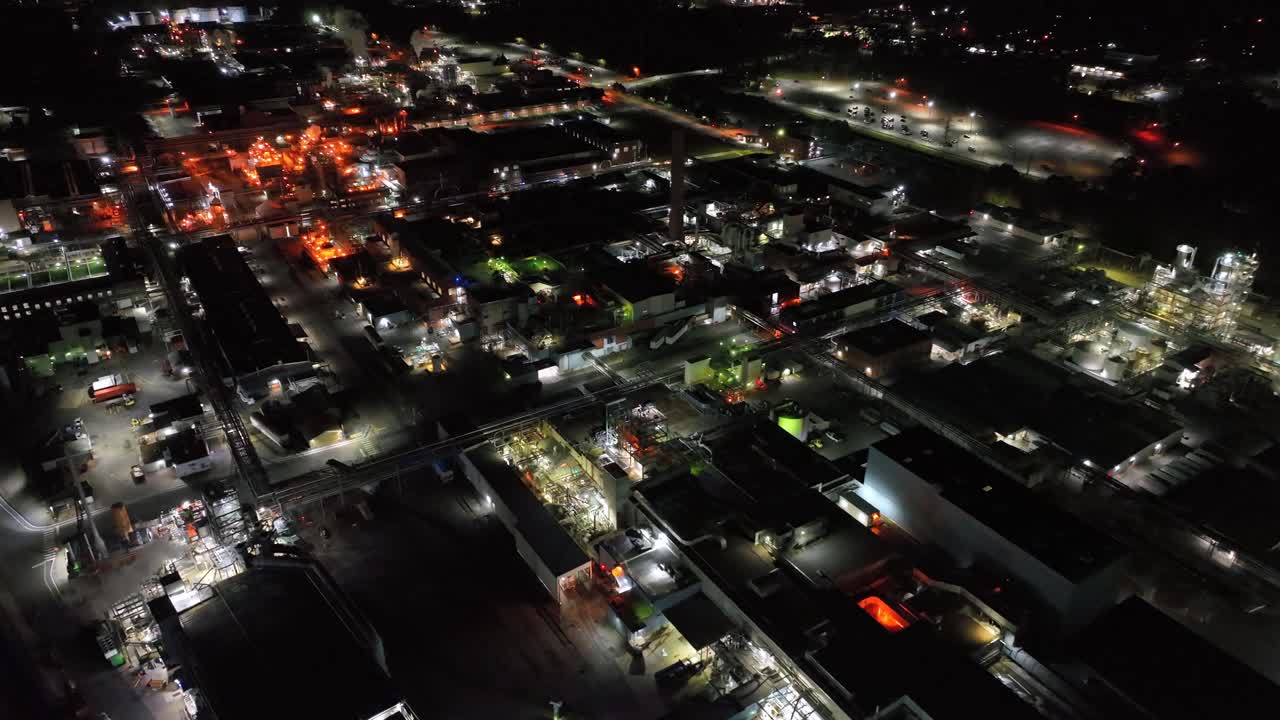 Panoramiche riprese aeree mostrano la Richmond DuPont Factory di notte, rivelando un complesso industriale incandescente, edifici illuminati, strade e luci della città su uno sfondo urbano scuro