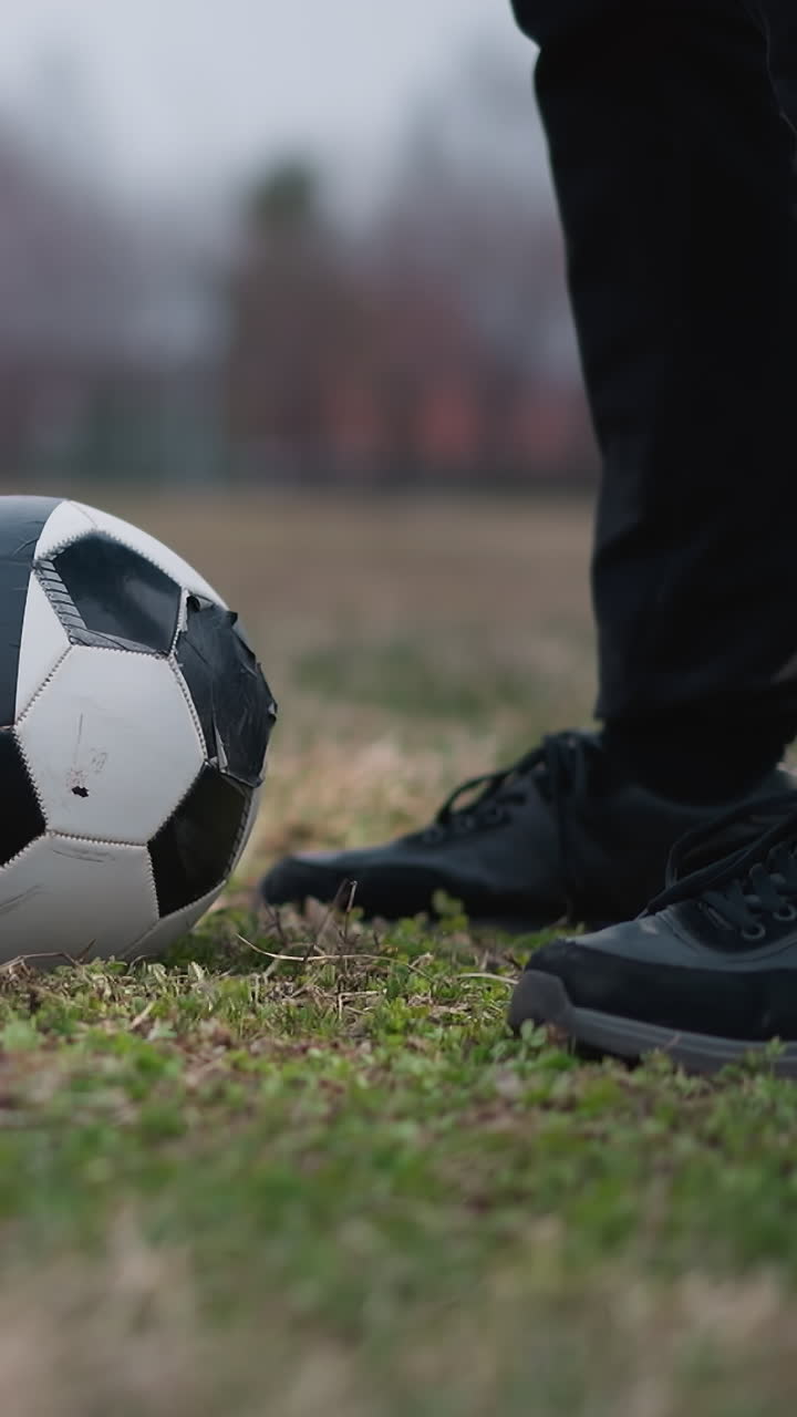 Close-up of a soccer ball on a grassy field, with a person standing nearby and another approaching close to the ball, the background is blurred, showing a football goalpost