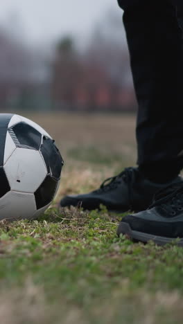 close-up of a soccer ball on a grassy field, with a person standing nearby and another approaching close to the ball, the background is blurred, showing a football goalpost