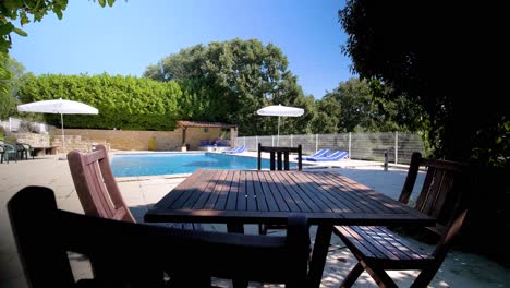 Slow-establishing-shot-of-a-wooden-table-sat-in-the-shade-beside-a-swimming-pool