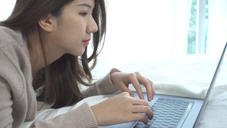 beautiful young smiling asian woman working on laptop while sitting on bed in bedroom at home. asian business woman relax in her home. enjoying time at home.