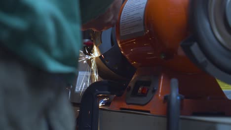 A-moving-low-angle-shot-of-a-mechanic-polishing-a-welded-metallic-car-piece,-with-sparks-flying-down-his-spinning-polishing-wheel