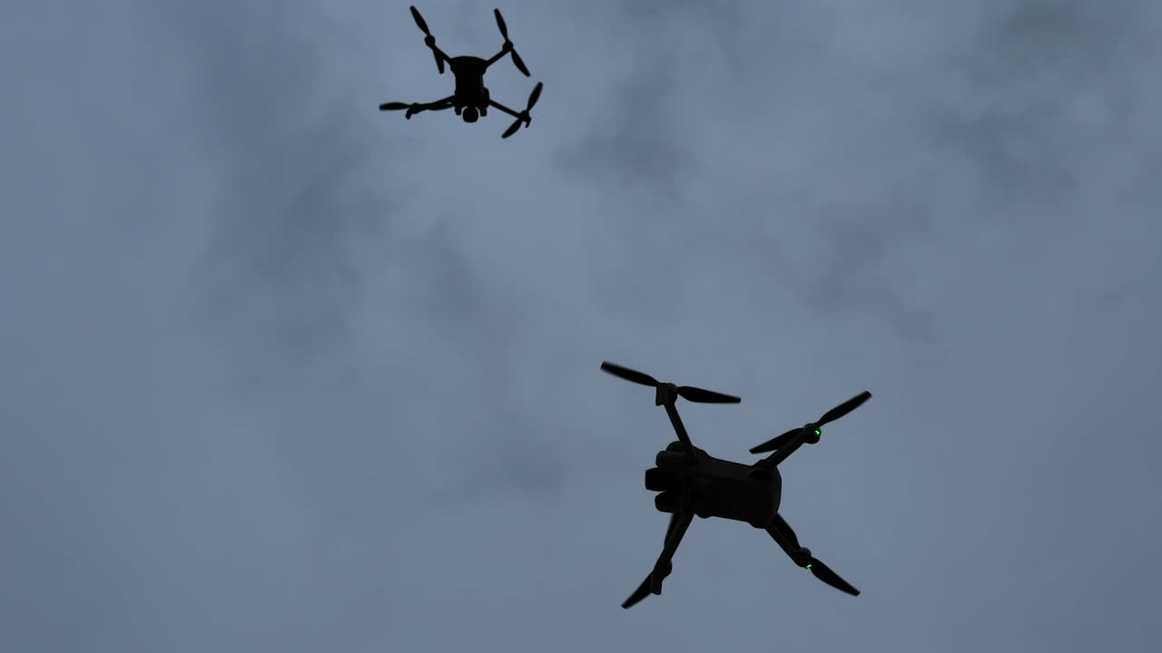To droner flyr på en skyet himmel over et åpent område på dagtid. Droner sees flygende på den skyete himmelen