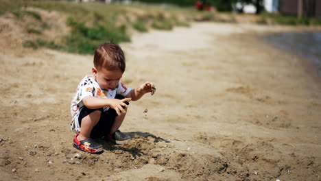 adorable caucasian toddler is focused on his game with the sand. kid sits on the river bank taking sand into hands. top view.