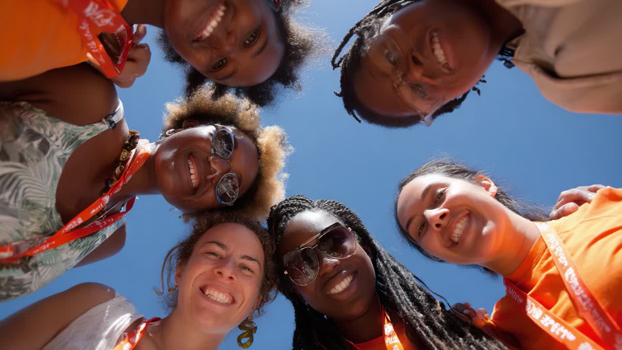 A group of diverse friends smiles brightly while standing in a huddle against a clear blue sky, showcasing their unity and joy in a vibrant and uplifting atmosphere