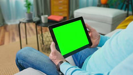close up of digital tablet with blank green mock up screen display hold by old adult man relaxing at home. camera moves away from the tablet