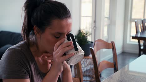 Medium-handheld-shot-of-a-pretty-millennial-caucasian-woman-who-slowly-slips-a-morning-cup-of-coffee-while-sitting-in-her-kitchen-and-dining-room