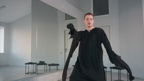 young man in flowing black costume dances expressively in front of large mirror in bright studio, gloved hands and dramatic movements emphasize fluid motion and moody artistic performance