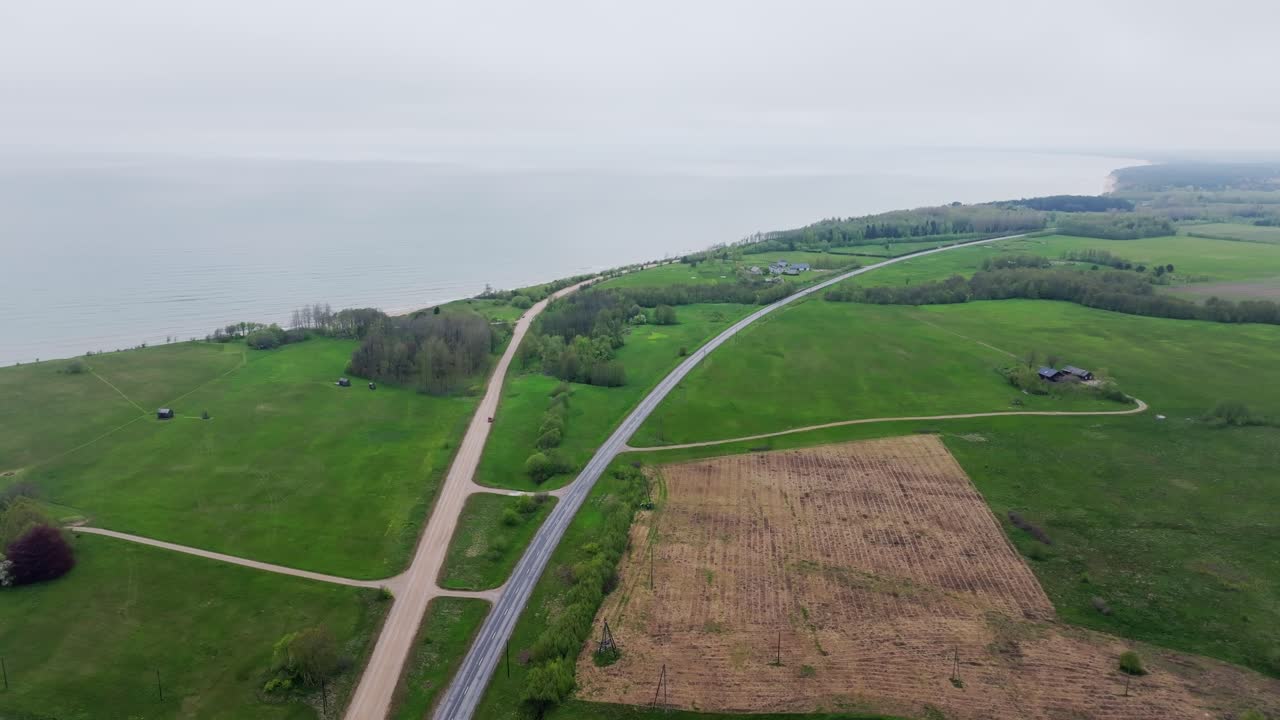 Drone flight over rural road leading along Latvia coast, Baltic Sea behind field