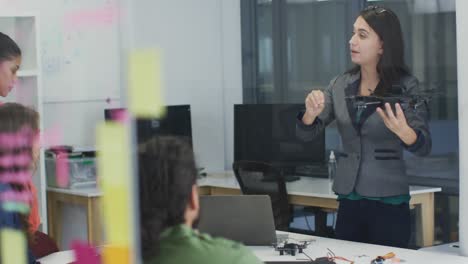 Mujer-Caucásica-Sosteniendo-Un-Dron-Dando-Una-Presentación-A-Un-Grupo-Diverso-De-Colegas