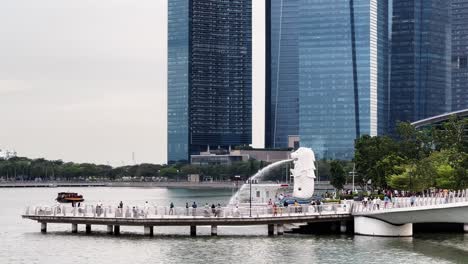 static shot of tourists and locals visiting the famous merlion park at downtown metropolitan area with business and financial buildings in the background and touristic river cruise sailing across