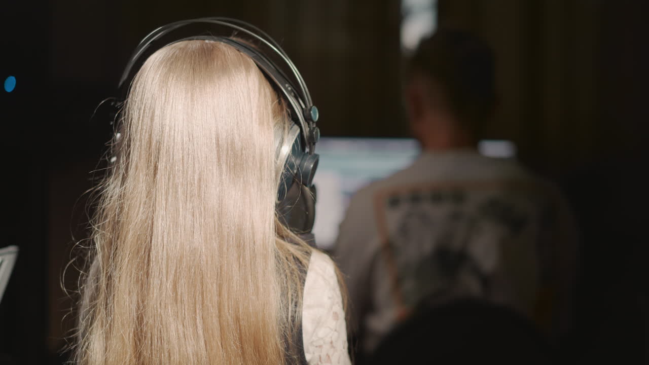 소녀는 교사와 함께 마이크에 노래를 부릅니다. 작은 금발 소녀는 녹음 스튜디오에서 보컬을 연습하는 동안 튜터 앞에 서 있습니다. 재능있는 소녀