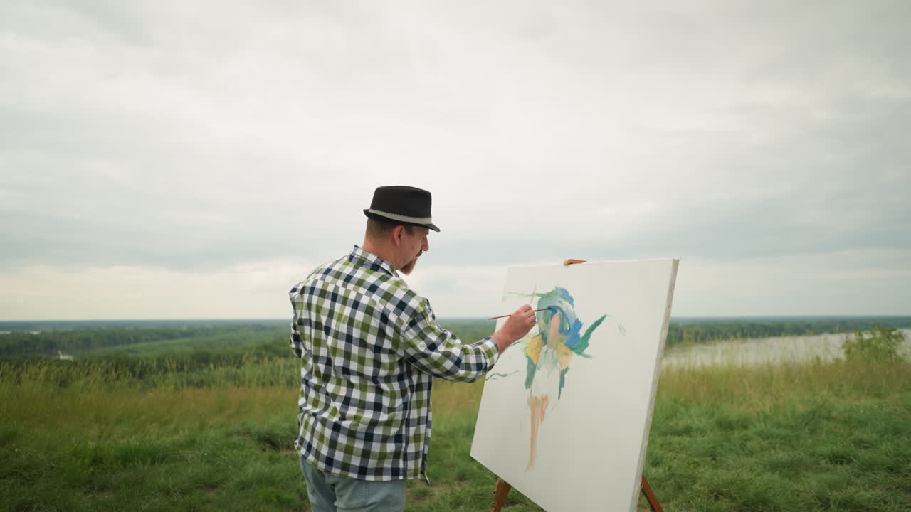 An artist, wearing a hat, checkered shirt, and jeans, is deeply focused on painting on a board in the middle of a lush grassy field under a cloudy sky