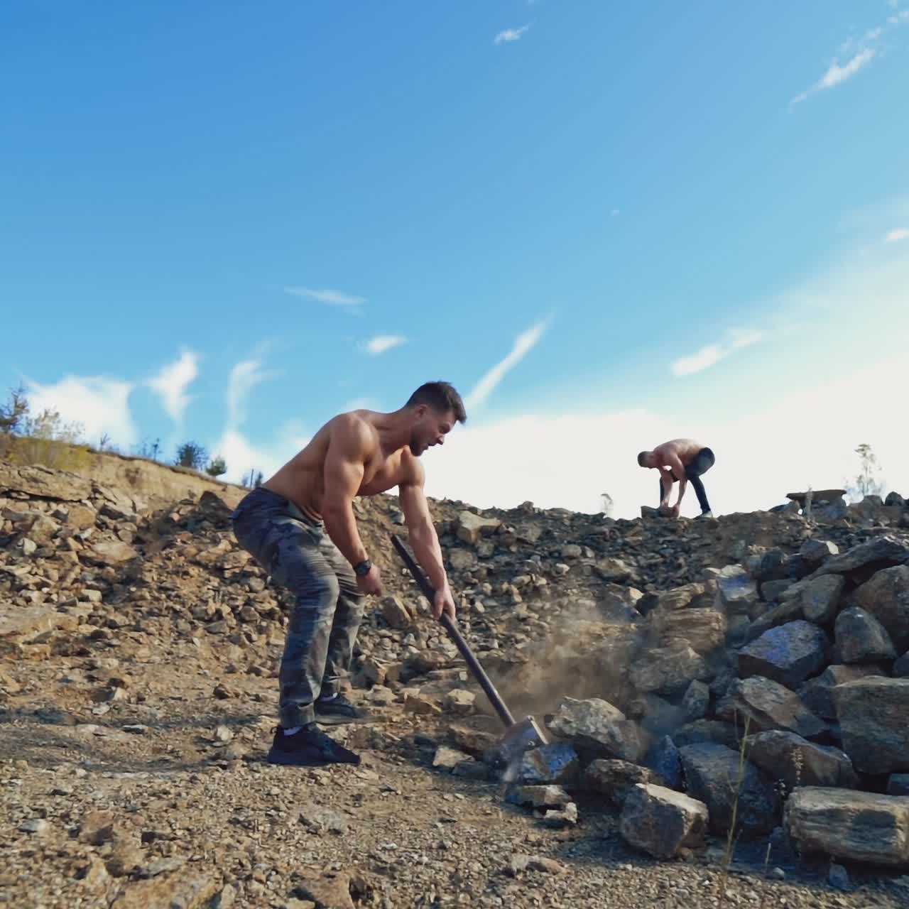 Strength sportsmen training on rocky background. One athlete breaking rocks with a heavy hammer while another man throwing stones. Slow motion.